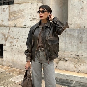 May include: A woman wearing a brown leather jacket with a distressed look, grey jeans, and brown sunglasses. She is holding a brown leather bag.