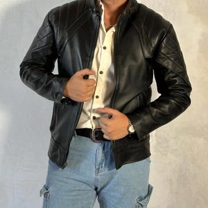 Puede incluir: Una chaqueta de cuero negra con un diseño acolchado. La chaqueta tiene una cremallera y un cuello alto. El modelo lleva una camisa blanca abotonada y unos vaqueros azules.