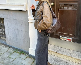 Op de afbeelding: Een vrouw draagt een bruin leren jack, een donkere jeans en zwarte enkellaarsjes. Ze draagt een bruine geweven handtas.