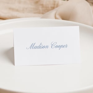 May include: A white rectangular folded place card featuring the name Madison Cooper printed in a blue cursive script font. The card is positioned upright on a smooth, matte white ceramic plate, displaying clear and legible typography.