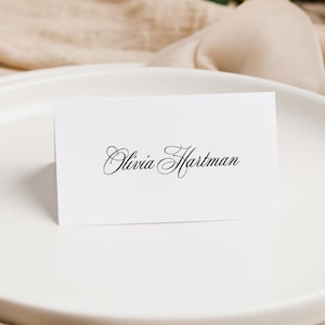 May include: A white rectangular folded place card featuring the name Olivia Hartman printed in a black elegant script font. The card is positioned upright on a smooth white ceramic plate, displaying clear black lettering against the white paper.