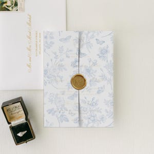 May include: A wedding invitation suite featuring a floral and butterfly pattern in light blue on white. The invitation is sealed with a gold wax seal. An envelope and a ring box are also present.