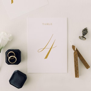 May include: A wedding table number card with the word "TABLE" and the number "4" in gold script. Two gold wedding rings are in a black velvet ring box. Also, there are two brown wax seals and a decorative stamp.