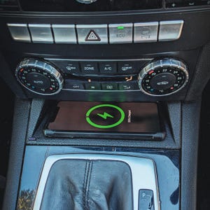 Puede incluir: Un smartphone cargando de forma inalámbrica en la consola central de un coche. El teléfono muestra un símbolo de carga verde y lee "95% Charged". El salpicadero del coche tiene mandos y botones de control climático.