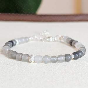 May include: A beaded bracelet featuring a gradient of gray and clear beads. The bracelet has a silver-colored chain and a decorative silver bead. The beads transition from light to dark gray.