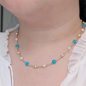 May include: A gold necklace featuring alternating turquoise and pearl beads, interspersed with textured gold accents. The necklace is displayed on a person wearing a white blouse.