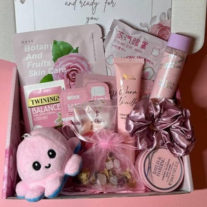 May include: A gift box filled with self-care items, including skincare products, tea, and a plush octopus. The box is white with a floral design and a card that reads "Moments of magic wrapped and ready for you". The items are mostly pink.