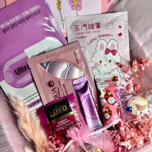 Op de afbeelding: Een geschenkdoos gevuld met schoonheidsproducten, waaronder gezichtsmaskers, oogmaskers, handcr&egrave;me en thee. De doos bevat een verscheidenheid aan roze en paarse verpakkingen, met tekst in meerdere talen. Een kaart met de tekst "Just a little thing to say" is ook inbegrepen.