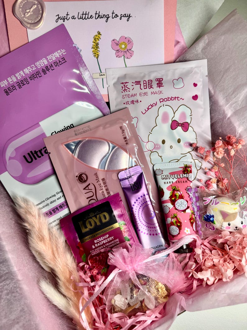 Op de afbeelding: Een geschenkdoos gevuld met schoonheidsproducten, waaronder gezichtsmaskers, oogmaskers, handcr&egrave;me en thee. De doos bevat een verscheidenheid aan roze en paarse verpakkingen, met tekst in meerdere talen. Een kaart met de tekst "Just a little thing to say" is ook inbegrepen.