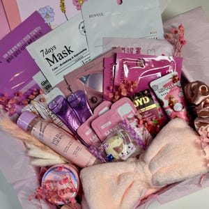 Op de afbeelding: Een roze geschenkdoos gevuld met schoonheidsproducten, waaronder gezichtsmaskers, lotions en een zachte haarband. De doos bevat diverse huidverzorgingsartikelen, decoratieve bloemen en een kaart met de tekst "Just a little thing to say."