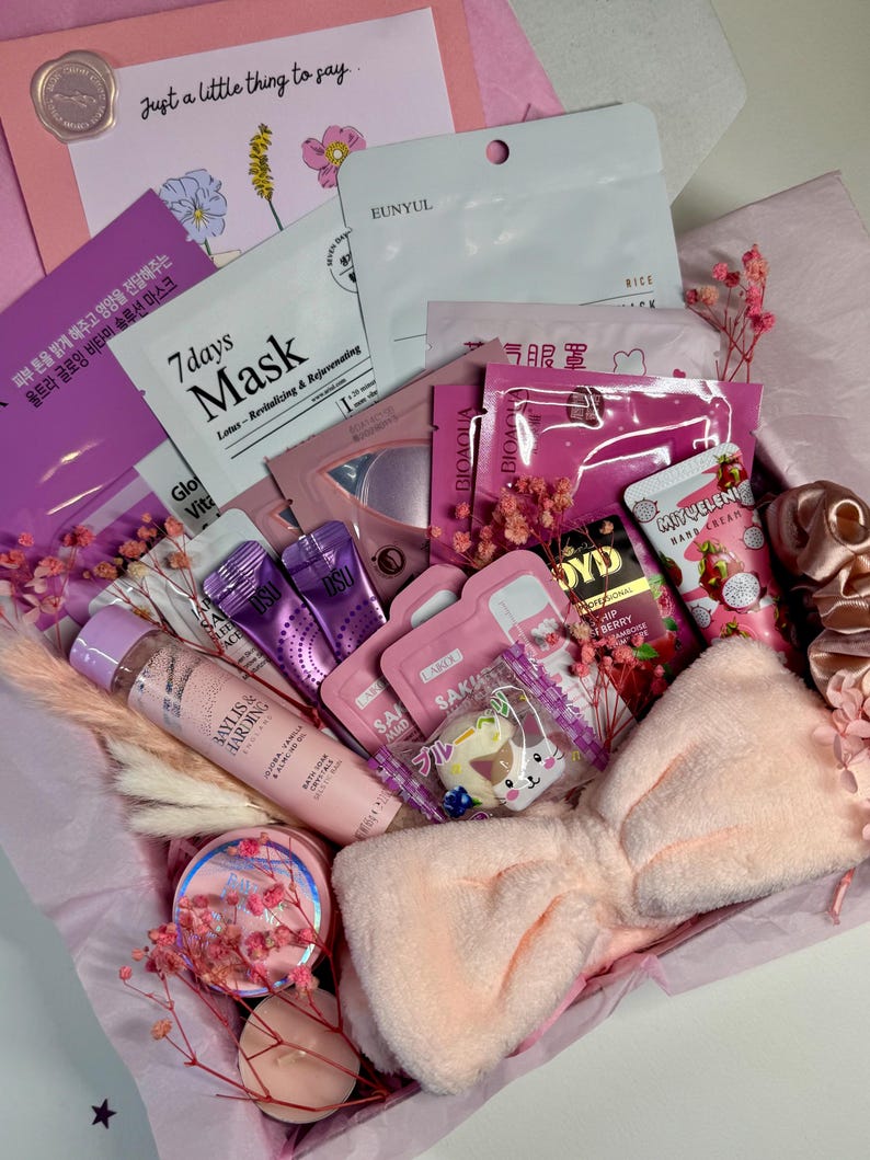 Op de afbeelding: Een roze geschenkdoos gevuld met schoonheidsproducten, waaronder gezichtsmaskers, lotions en een zachte haarband. De doos bevat diverse huidverzorgingsartikelen, decoratieve bloemen en een kaart met de tekst "Just a little thing to say."