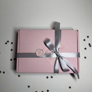 Op de afbeelding: Een roze rechthoekige geschenkdoos vastgebonden met een zilveren lint en strik. Een cirkelvormige waszegel is op de doos aangebracht. De doos staat op een wit oppervlak bezaaid met zwarte stervormige confetti.