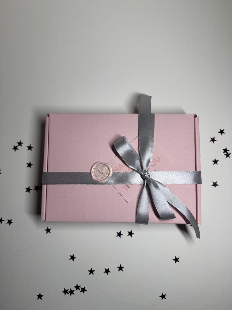 Op de afbeelding: Een roze rechthoekige geschenkdoos vastgebonden met een zilveren lint en strik. Een cirkelvormige waszegel is op de doos aangebracht. De doos staat op een wit oppervlak bezaaid met zwarte stervormige confetti.
