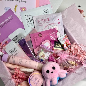 Op de afbeelding: Een roze geschenkdoos gevuld met schoonheidsproducten, waaronder gezichtsmaskers, lotions en een pluche octopus. De doos bevat items met tekst in meerdere talen en een kaart met de tekst "Just a little thing to say."