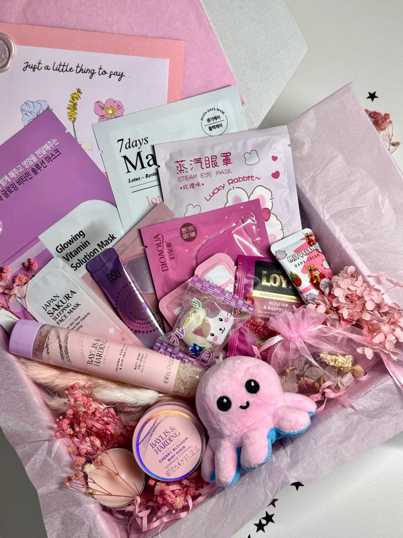 Op de afbeelding: Een roze geschenkdoos gevuld met schoonheidsproducten, waaronder gezichtsmaskers, lotions en een pluche octopus. De doos bevat items met tekst in meerdere talen en een kaart met de tekst "Just a little thing to say."