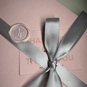 Op de afbeelding: Een roze geschenkdoos, vastgebonden met een grijs satijnen lint, met een strik en een zegel. De zegel heeft tekst en een ontwerp. Een kaart met tekst is zichtbaar onder het lint.