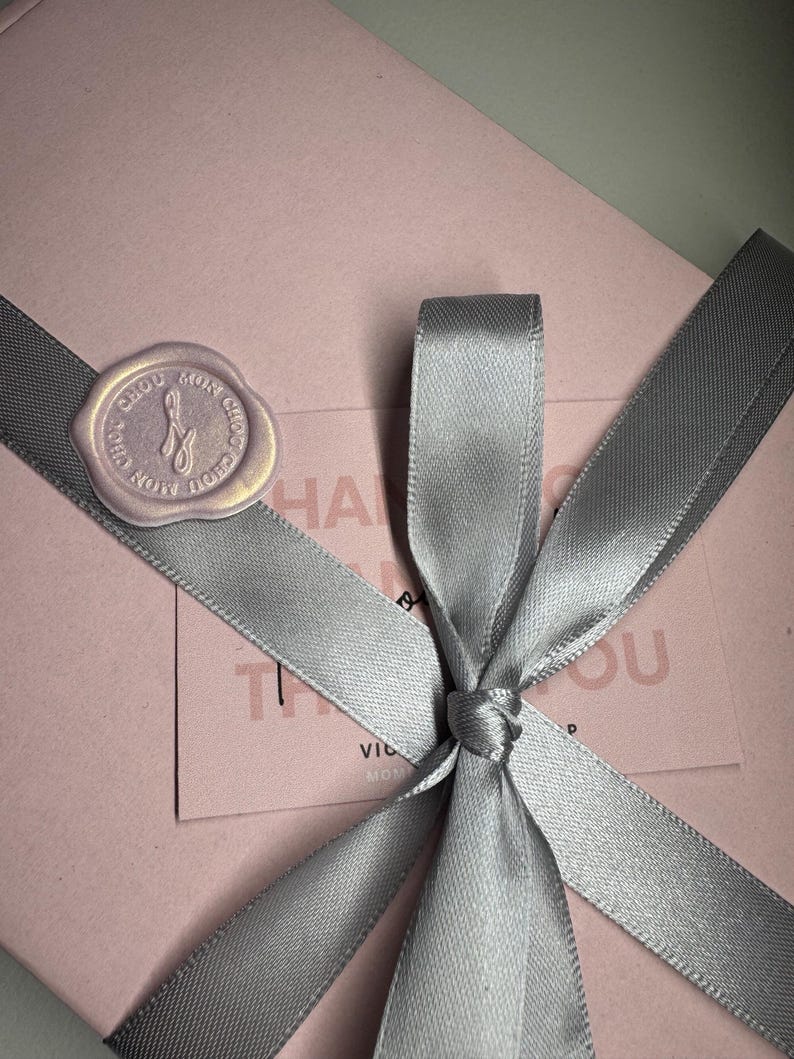 Op de afbeelding: Een roze geschenkdoos, vastgebonden met een grijs satijnen lint, met een strik en een zegel. De zegel heeft tekst en een ontwerp. Een kaart met tekst is zichtbaar onder het lint.