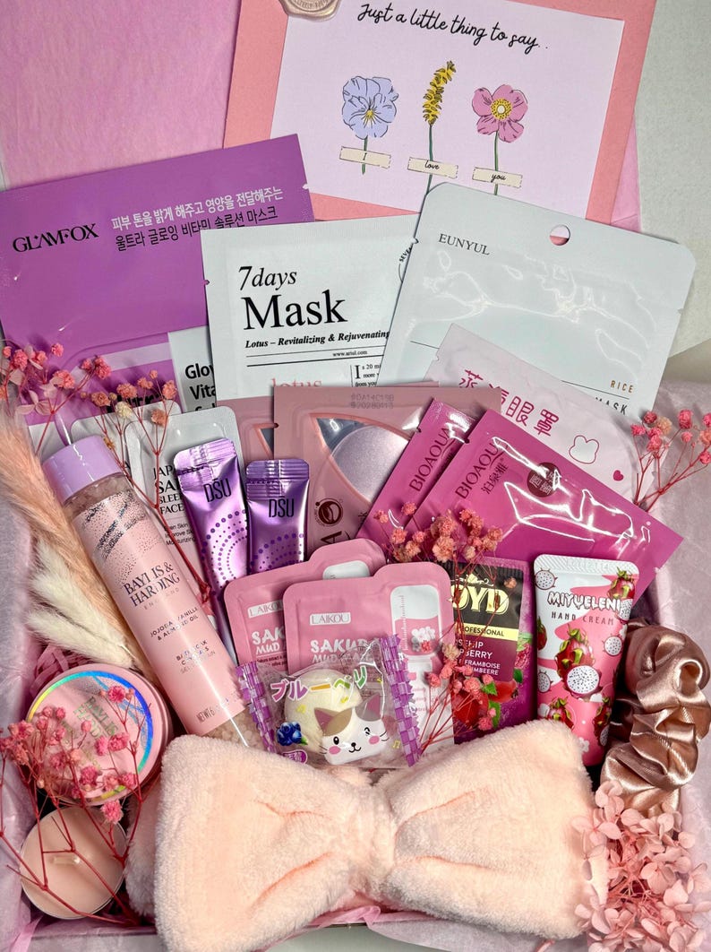 Op de afbeelding: Een roze geschenkdoos gevuld met schoonheidsproducten, waaronder gezichtsmaskers, huidverzorgingsartikelen en een pluche strik. De doos bevat items met tekst in meerdere talen en decoratieve droogbloemen.