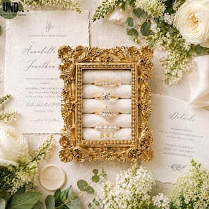 May include: A rectangular gold ornate frame ring holder with a cream velvet interior featuring four horizontal slots holding four rings and the engraved letters B heart L, surrounded by white flowers and wedding invitation stationery.