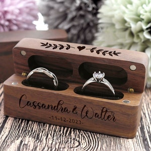 K&ouml;nnte beinhalten: Eine Holzkiste f&uuml;r Ringe mit zwei F&auml;chern f&uuml;r Ringe. Die Kiste ist mit den Namen "Cassandra & Walter" und dem Datum "11-12-2023" graviert. Die Oberseite der Kiste ist mit einem Herz und Bl&auml;ttern verziert.
