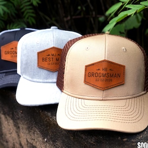 Może przedstawiać: Trzy spersonalizowane czapki typu trucker z naszywkami ze sztucznej skóry. Czapki są w kolorze czarnym, szarym i beżowym, każda z innym imieniem i datą. Naszywki głoszą "Groom", "Best Man" i "Groomsman". Czapki mają siatkową tylną część i zakrzywiony daszek.