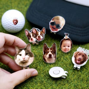 May include: A collection of personalized golf ball markers and a golf ball, each featuring a custom image of a pet or person. The markers are attached to a magnetic clip. The golf ball has a small image of a dog wearing a crown.