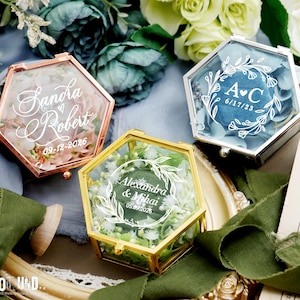 Op de afbeelding: Drie zeshoekige ringdozen in roségoud, goud en zilver, elk met een glazen bovenkant en gevuld met bloemen. De dozen hebben gepersonaliseerde tekst met namen en data, perfect voor bruiloften of verlovingen.