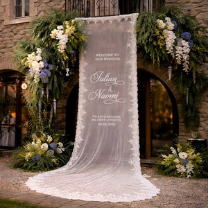 May include: A wedding ceremony entrance decorated with greenery and white and blue flowers. A sheer white fabric banner with lace trim displays the names "Julian & Naomi" and the date 14.06.2026, with the text "WELCOME TO OUR WEDDING" above.