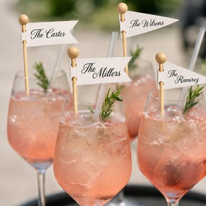 Op de afbeelding: Vier doorzichtige wijnglazen gevuld met een roze cocktail, ijs en een takje rozemarijn. Elk glas bevat een houten roerstaafje met een klein wit wimpelvlaggetje waarop de namen The Carter, The Millers, The Wilsons en The Ramirez staan gedrukt.