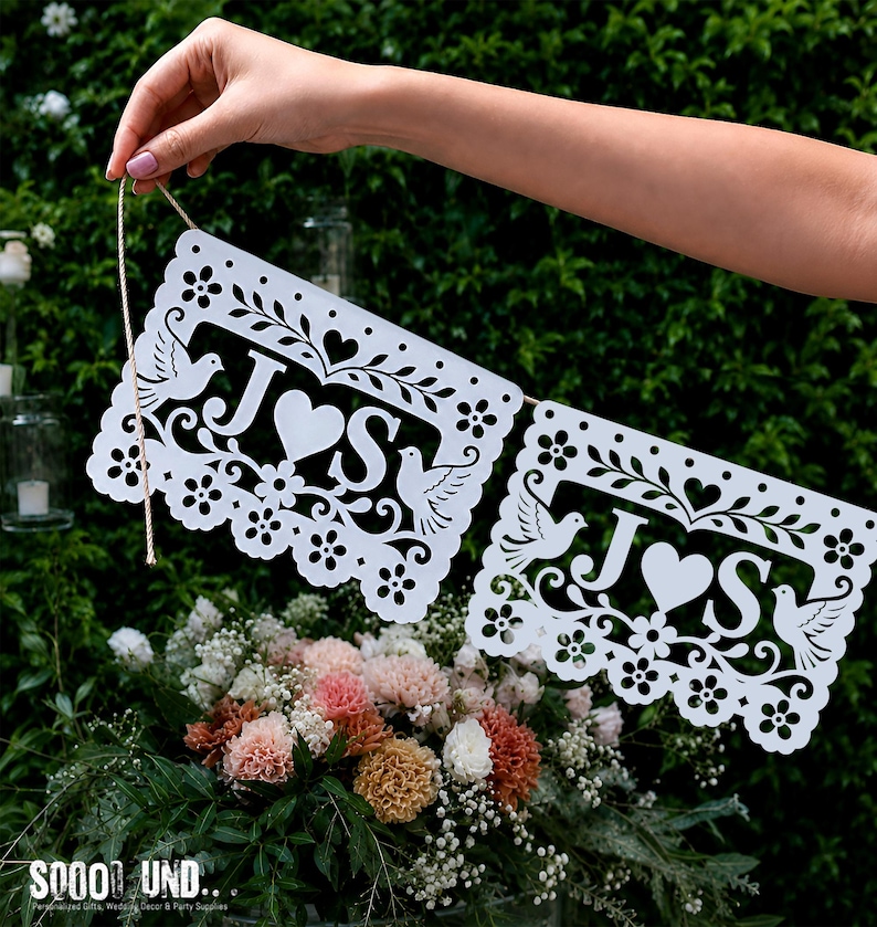 May include: Two white rectangular paper banners featuring intricate laser-cut designs of doves, flowers, and hearts. Each banner displays the letters J and S separated by a central heart. The banners are connected by a thin, light brown twine string.