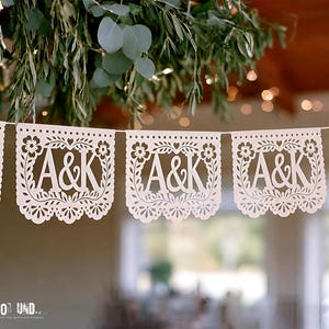 May include: A white paper banner featuring repeating cut-out panels with the letters A&K centered within a decorative floral border. Each rectangular panel has a scalloped bottom edge and is strung together on a thin white cord for display.