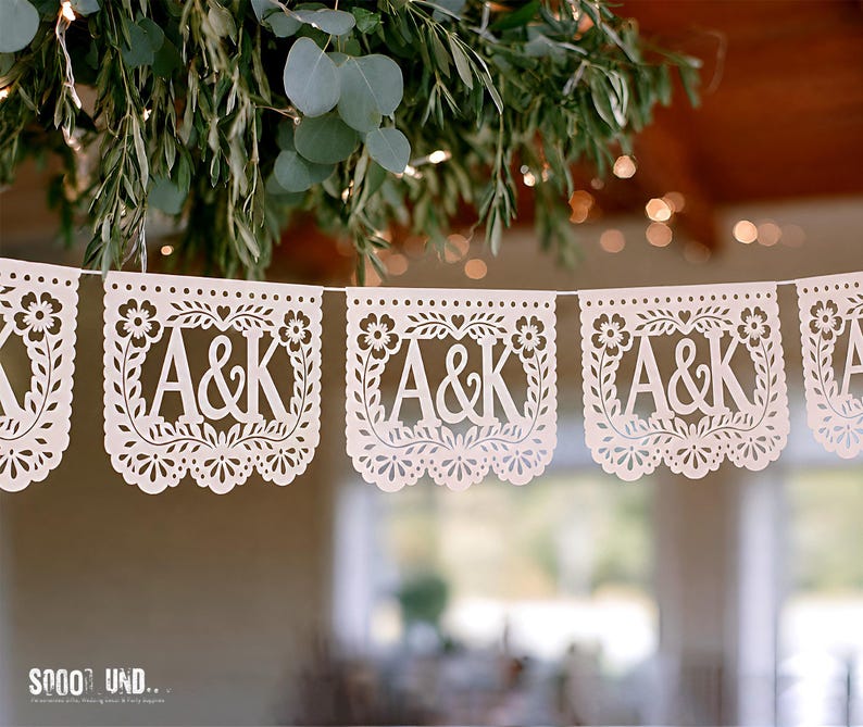 May include: A white paper banner featuring repeating cut-out panels with the letters A&K centered within a decorative floral border. Each rectangular panel has a scalloped bottom edge and is strung together on a thin white cord for display.