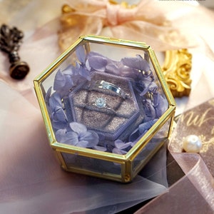 May include: A hexagonal glass box with a gold trim, containing a purple velvet ring box with two silver rings and a single diamond ring. The box is surrounded by dried lavender flowers.