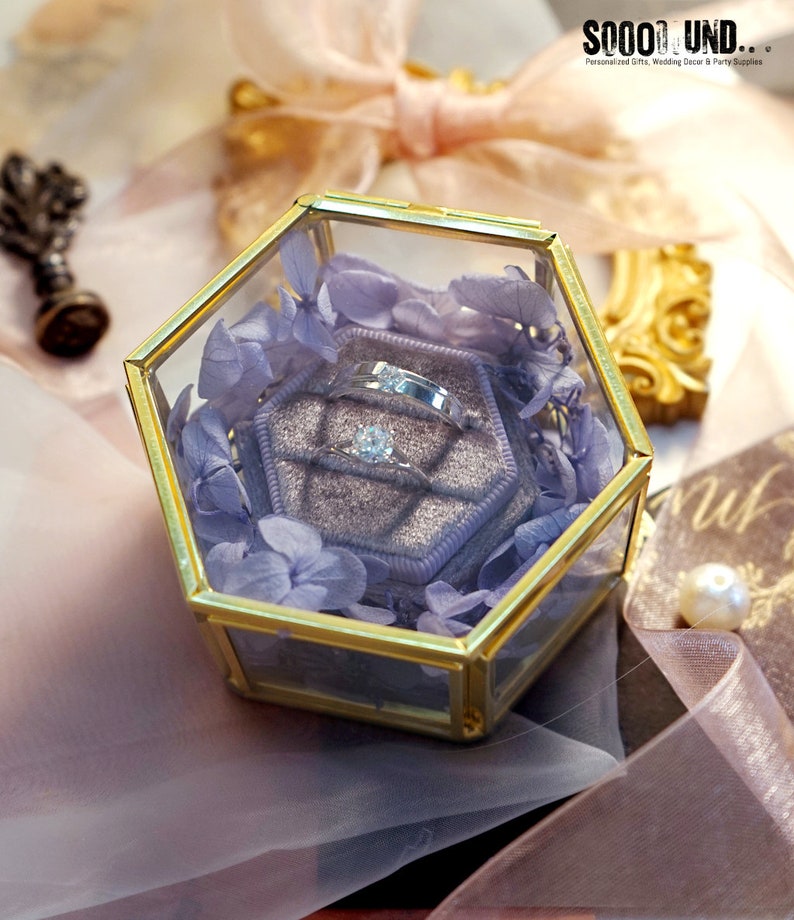 May include: A hexagonal glass box with a gold trim, containing a purple velvet ring box with two silver rings and a single diamond ring. The box is surrounded by dried lavender flowers.