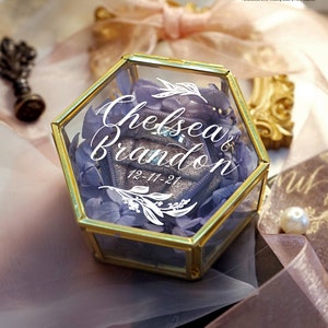 May include: A gold hexagonal glass ring box with a purple flower arrangement inside. The box has the names "Chelsea & Brandon" and the date "12-11-21" written on it.