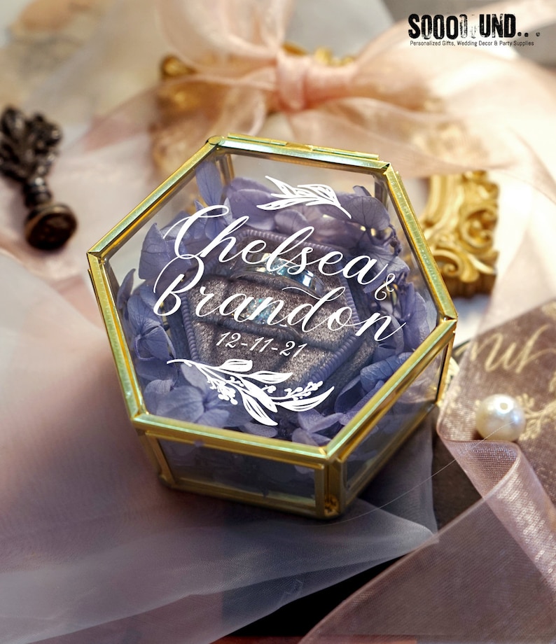 May include: A gold hexagonal glass ring box with a purple flower arrangement inside. The box has the names "Chelsea & Brandon" and the date "12-11-21" written on it.