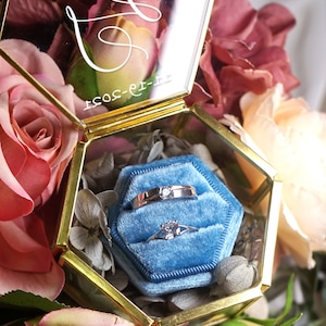May include: A gold hexagonal glass box with a blue velvet interior. The box contains two silver rings, one with a single diamond. The box is surrounded by pink and red roses.