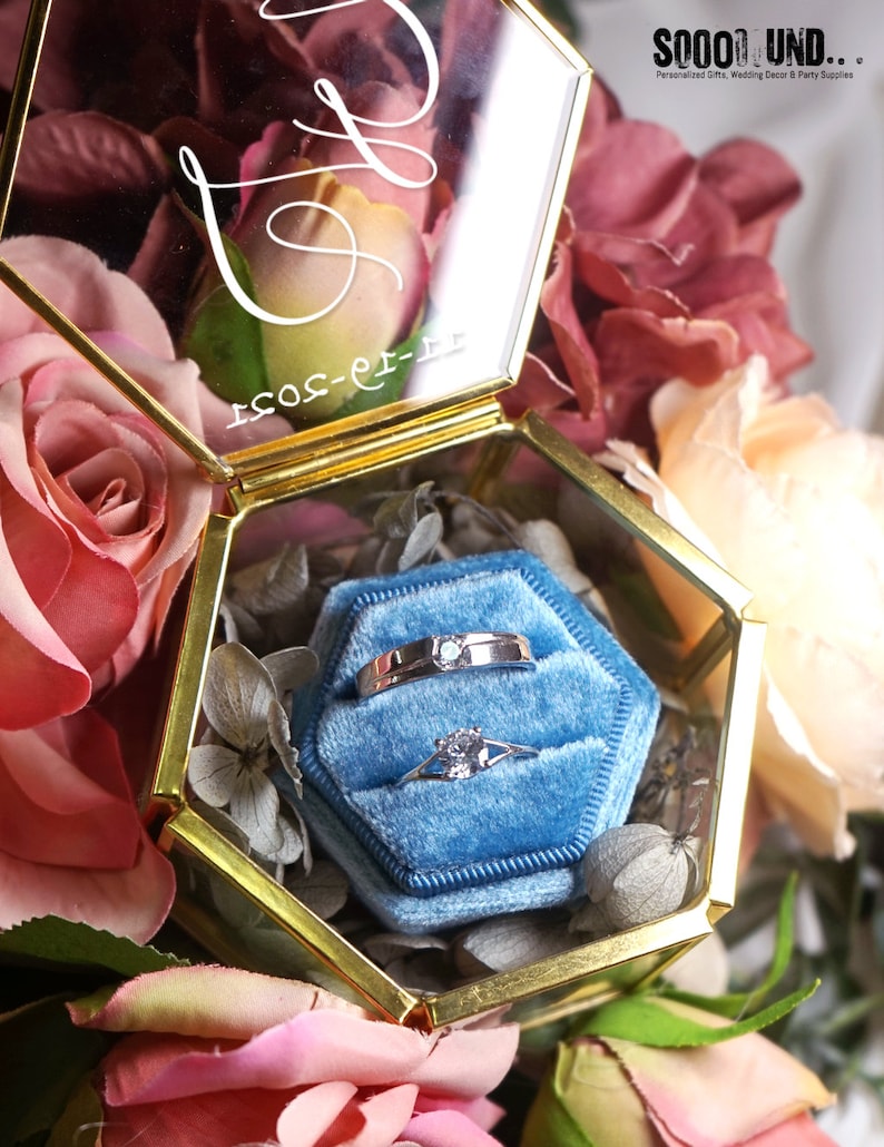 May include: A gold hexagonal glass box with a blue velvet interior. The box contains two silver rings, one with a single diamond. The box is surrounded by pink and red roses.