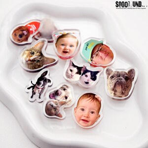 May include: A collection of clear acrylic personalized photo magnets featuring images of pets, babies, and people. The magnets are in various shapes and sizes, with a white border. The background is a white, curved tray. The text "SOOOO SOUND..." is visible in the top right corner.