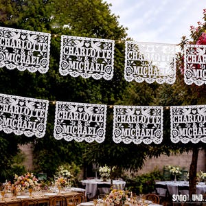 May include: A series of white rectangular paper banners hanging on a string, each featuring intricate laser-cut designs of two doves, a heart, and floral patterns. The text "CHARLOTTE & MICHAEL" is clearly cut out in the center of each banner.