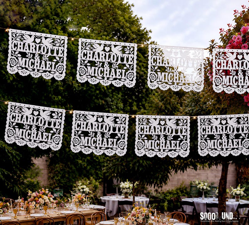 May include: A series of white rectangular paper banners hanging on a string, each featuring intricate laser-cut designs of two doves, a heart, and floral patterns. The text "CHARLOTTE & MICHAEL" is clearly cut out in the center of each banner.