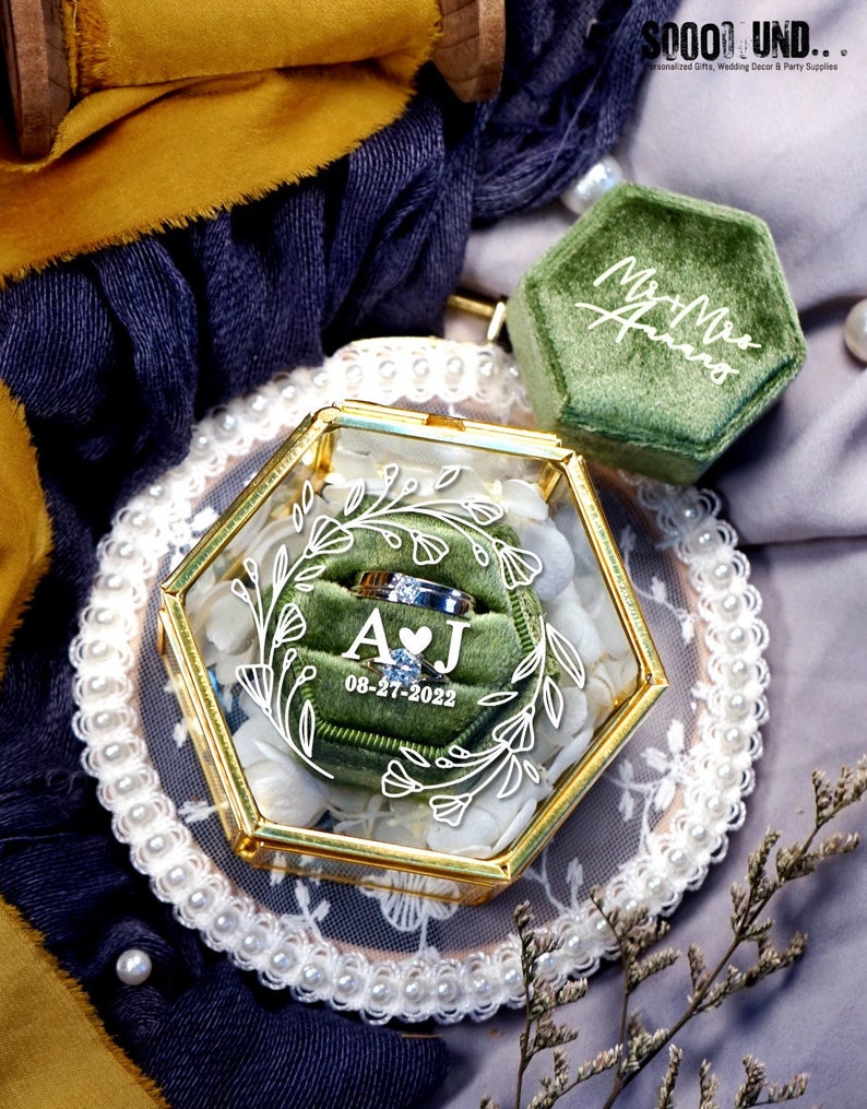 May include: A hexagonal glass ring box with a gold trim and a green velvet interior. The box has a floral design and the initials "AJ" with a heart and the date "08-27-2022".