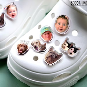 May include: White clogs adorned with various clear, personalized charms. The charms feature images of a baby, a man, dogs, and cats. The charms are attached to the top of the clogs. The text "SOOOO UND" is visible in the upper right corner.
