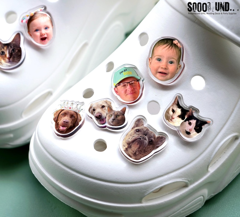 May include: White clogs adorned with various clear, personalized charms. The charms feature images of a baby, a man, dogs, and cats. The charms are attached to the top of the clogs. The text "SOOOO UND" is visible in the upper right corner.