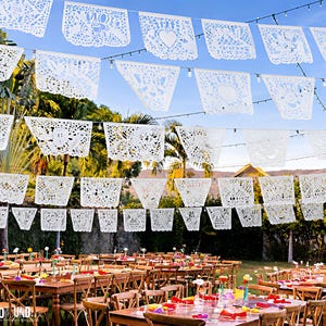 May include: Multiple rows of white rectangular paper banners with intricate laser-cut designs featuring hearts and floral patterns, suspended on strings above several wooden tables arranged in an outdoor setting with palm trees in the background.