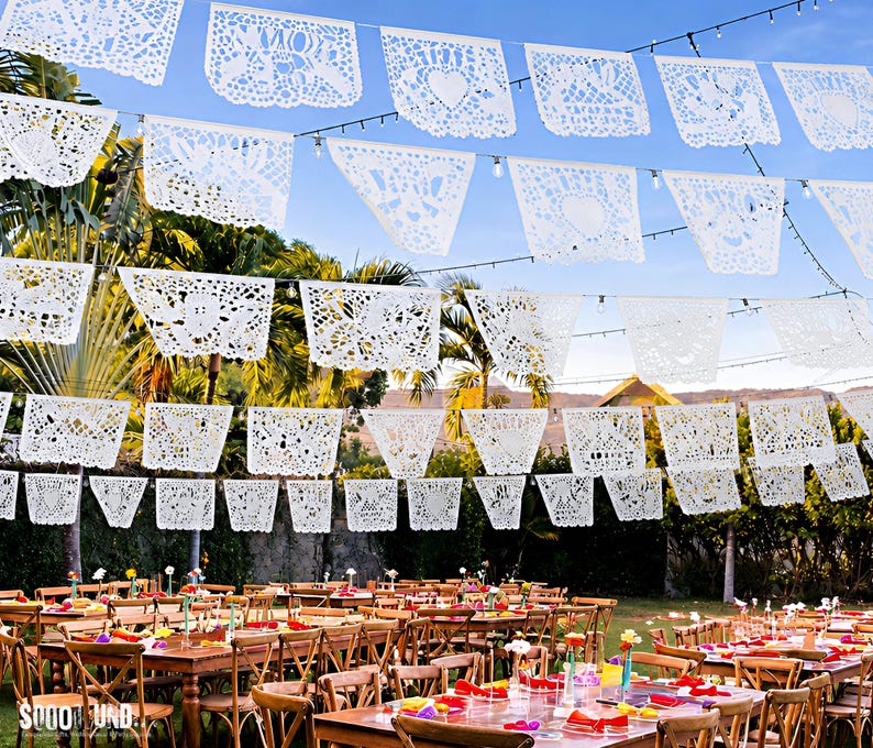 May include: Multiple rows of white rectangular paper banners with intricate laser-cut designs featuring hearts and floral patterns, suspended on strings above several wooden tables arranged in an outdoor setting with palm trees in the background.