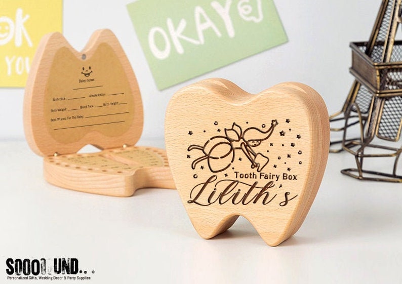 Puede incluir: Una caja de dientes de hada de madera con forma de diente con un dise&ntilde;o de hada y el texto "Tooth Fairy Box Lilith's". La caja tiene una tapa con bisagras y un peque&ntilde;o compartimento en el interior para guardar los dientes perdidos.