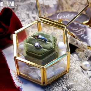 May include: A gold hexagonal glass ring box with a green velvet interior. Two silver rings with diamonds are displayed inside the box.