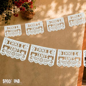 May include: A white paper banner featuring multiple rectangular panels strung together. Each panel displays the text EMMA & JAMES in a bold font, surrounded by intricate floral and leaf cut-out patterns along the edges and borders.