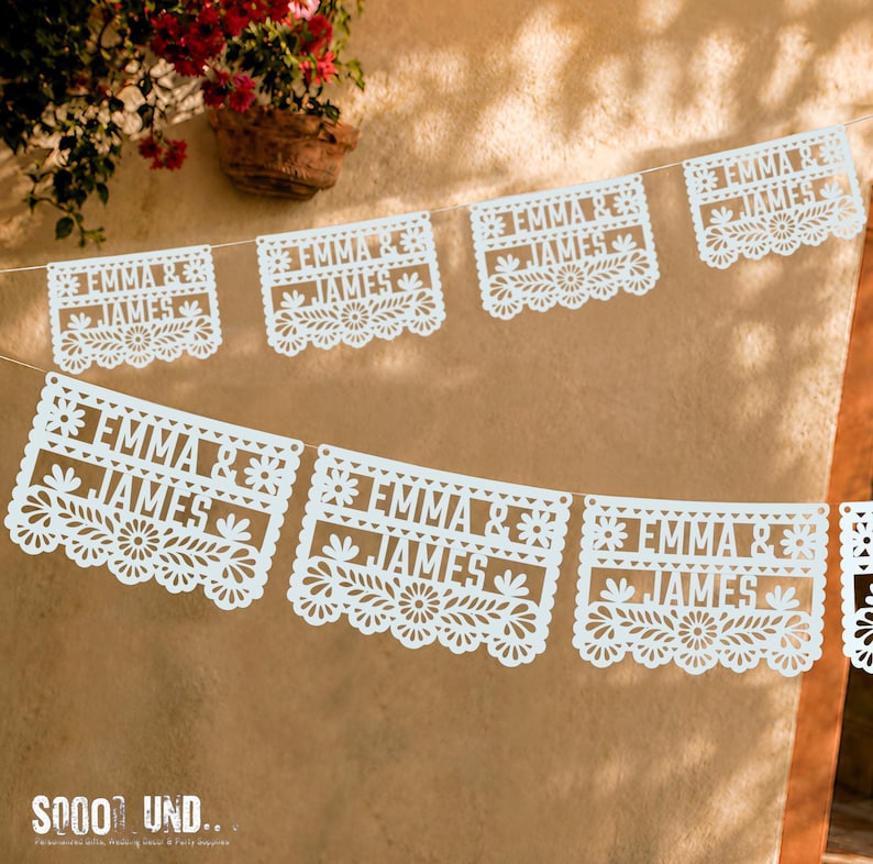 May include: A white paper banner featuring multiple rectangular panels strung together. Each panel displays the text EMMA & JAMES in a bold font, surrounded by intricate floral and leaf cut-out patterns along the edges and borders.